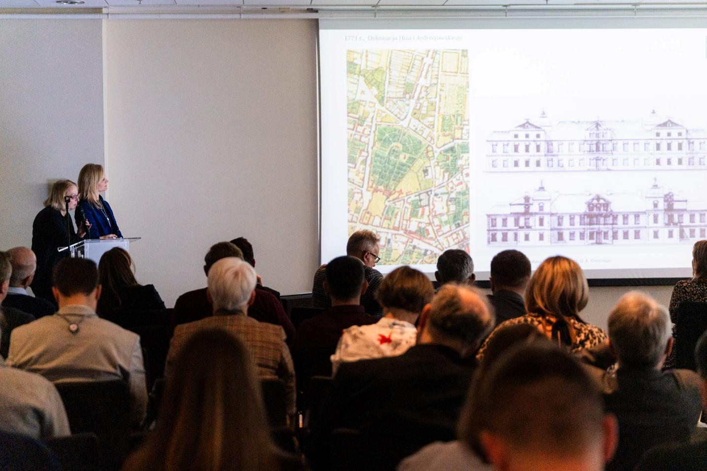 Na pierwszym planie zdjęcia odwróceni tyłem uczestnicy konferencji, za nimi po lewej stronie przy pulpicie z mikrofonem dwie prelegentki. Po prawej stronie ekran rzutnika z wyświetloną mapą fragmentu Warszawy (z Ogrodem Saskim) oraz szkicami Pałacu Brühla, dokumentującymi przemiany jego wyglądu.